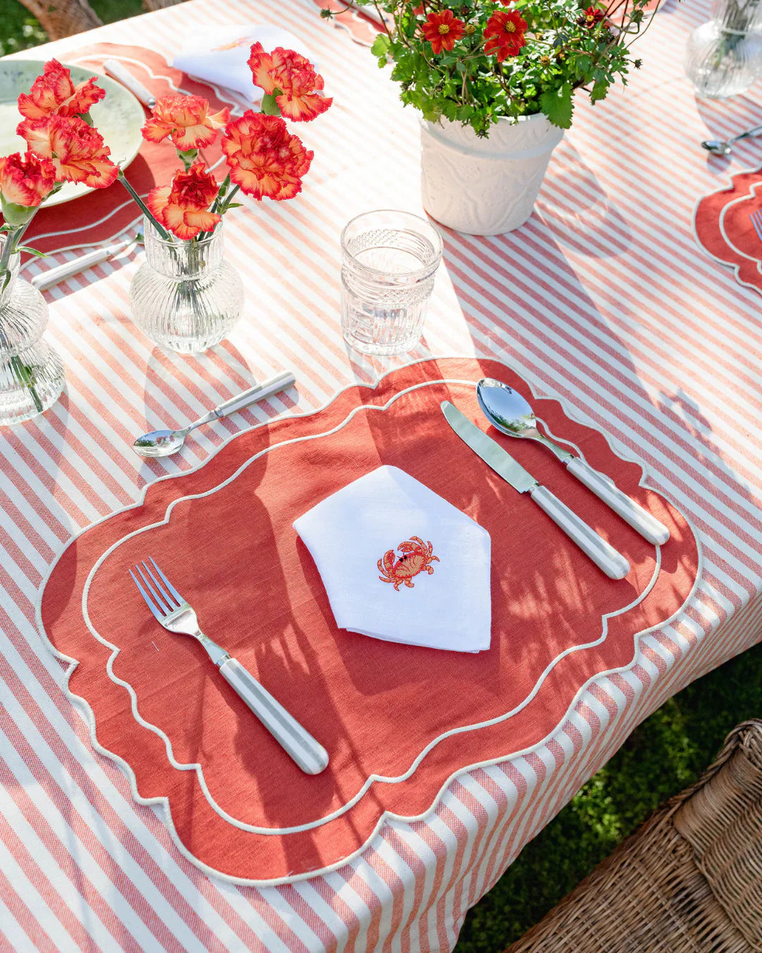 Disposición elegante de cubiertos, cristalería y servilleta blanca bordada sobre un mantel individual rojo en una mesa al aire libre, ilustrando el protocolo en la mesa.